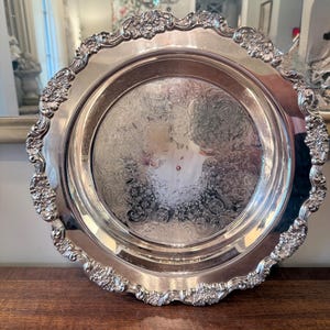 Puede incluir: Una bandeja de servicio redonda de metal plateado con un borde floral en relieve adornado y bordes festoneados. El centro de la bandeja muestra un intrincado patrón de volutas grabadas que cubre toda la superficie plana del plato.
