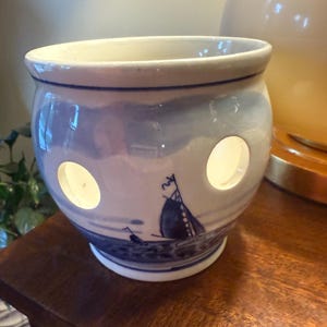 May include: A white ceramic planter with a blue and white Delftware design featuring a sailboat. The planter has three drainage holes.