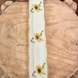 May include: A long, narrow, rectangular porcelain tray with wavy edges and a gold-trimmed rim. The white surface features three hand-painted yellow sunflower motifs spaced evenly along the center of the dish.