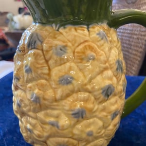 May include: A ceramic pineapple pitcher with a green top and yellow body. The pitcher has a handle and is decorated with blue and brown details.