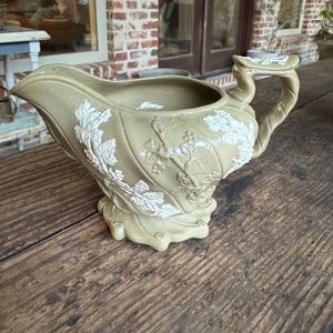 May include: A ceramic gravy boat with a white floral design. The boat is a light brown color and has a handle on the side.