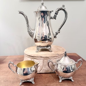 Op de afbeelding: Een zilveren theeservies met sierlijke details. De set bevat een hoge theepot, een creamer met een gouden interieur en een suikerpot met deksel. De stukken worden gepresenteerd op een houten oppervlak.
