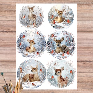 May include: Watercolour illustrations of deer and birds in a winter scene. Each circular design features a deer surrounded by snow-covered branches and small red birds. The artwork is on a white sheet with scissors and paintbrushes nearby.