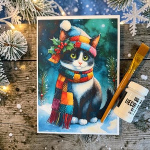 May include: A rectangular print featuring a black and white cat wearing a multicoloured patchwork hat and scarf with holly berries, positioned next to a wooden paintbrush and a small white bottle of decoupage glue on a rustic wooden surface.