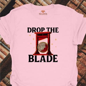 May include: A light pink t-shirt featuring a graphic of a red guillotine with a disco ball in place of the blade. The text "DROP THE BLADE" is printed in black above and below the illustration. Small yellow star icons surround the guillotine.