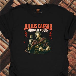 May include: A black short-sleeved t-shirt featuring a graphic of Julius Caesar holding a microphone. The text reads JULIUS CAESAR WORLD TOUR in orange and white. The design includes red lightning bolts and a small logo at the neckline.