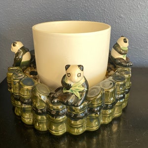 May include: A ceramic planter with a cream-colored pot and a decorative base featuring panda bear figurines. The base is designed to resemble bamboo stalks, with small pebbles around the pot's base.