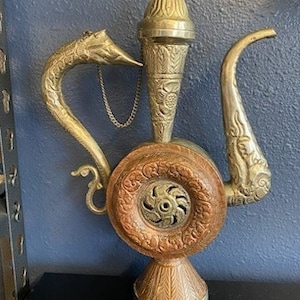 May include: An ornate, antique metal teapot with a dragon-shaped spout and decorative accents. The teapot features a circular body with a central design and a conical top. The colors are gold and brown.