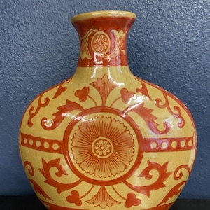 May include: A decorative ceramic vase with a rounded body and a narrow neck. The vase features a yellow base color with intricate red floral patterns and designs. The rim and base of the vase are also red.