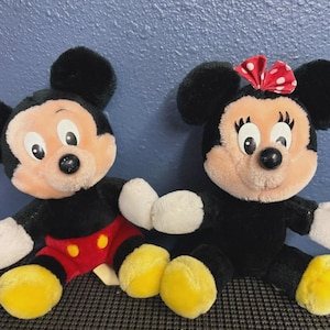 May include: Two plush Disney character dolls, Mickey Mouse and Minnie Mouse, are seated side-by-side. Mickey wears red shorts and yellow shoes. Minnie wears a red polka-dot bow and yellow shoes. Both have black fur, white gloves, and peach-colored faces.