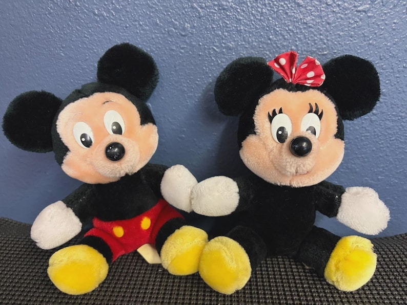 May include: Two plush Disney character dolls, Mickey Mouse and Minnie Mouse, are seated side-by-side. Mickey wears red shorts and yellow shoes. Minnie wears a red polka-dot bow and yellow shoes. Both have black fur, white gloves, and peach-colored faces.