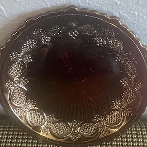 May include: A round, dark amber-colored glass plate with a decorative, embossed floral and geometric pattern around the rim. The plate has a scalloped edge and a smooth, reflective surface.