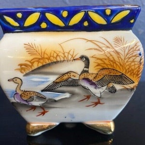 May include: A small, ceramic planter with a hand-painted design of ducks in a marsh setting. The planter has a white body, a blue rim with yellow accents, and gold-colored feet. The ducks are depicted with brown, purple, and white feathers.