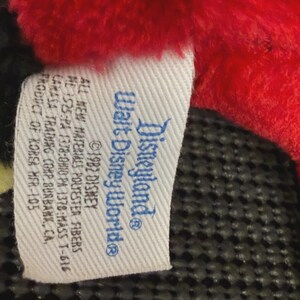 May include: A close-up of a tag from a Disneyland/Walt Disney World plush toy. The tag is white with blue text and features the Disneyland and Walt Disney World logos. The tag also includes text about the materials and manufacturer.