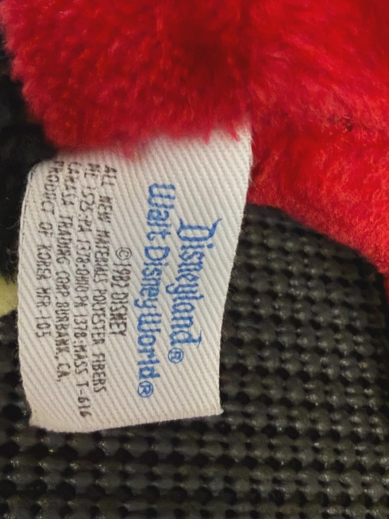 May include: A close-up of a tag from a Disneyland/Walt Disney World plush toy. The tag is white with blue text and features the Disneyland and Walt Disney World logos. The tag also includes text about the materials and manufacturer.