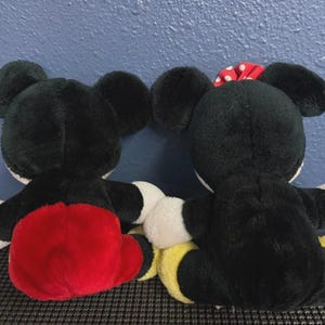 May include: Two plush Mickey Mouse and Minnie Mouse dolls, viewed from the back. Mickey wears red shorts, while Minnie has a red polka dot bow. Both have black fur, white gloves, and yellow shoes.