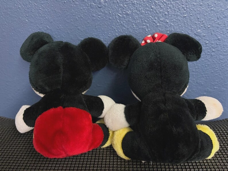 May include: Two plush Mickey Mouse and Minnie Mouse dolls, viewed from the back. Mickey wears red shorts, while Minnie has a red polka dot bow. Both have black fur, white gloves, and yellow shoes.