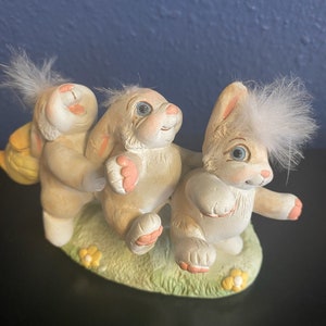 May include: A ceramic figurine featuring three light brown bunnies with pink paws and ears, and fluffy white accents. The bunnies are posed on a green base with yellow flowers, creating a whimsical Easter theme.