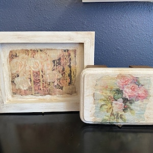 May include: A white-framed picture and a small box with a floral design. The picture frame has a distressed white finish and a floral print. The box has a similar distressed white finish and a floral print with pink roses.