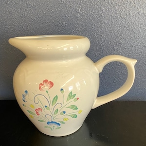 May include: Cream-colored ceramic pitcher with a floral design. The pitcher has a rounded body, a spout, and a curved handle. The floral design features red, blue, and yellow flowers with green leaves. The pitcher is likely used for serving beverages.