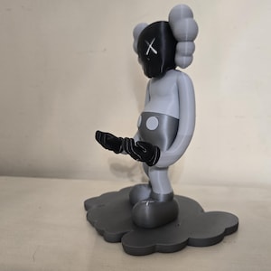 May include: A gray and black figurine of a character with a black X on its face, standing on a gray cloud-shaped base.