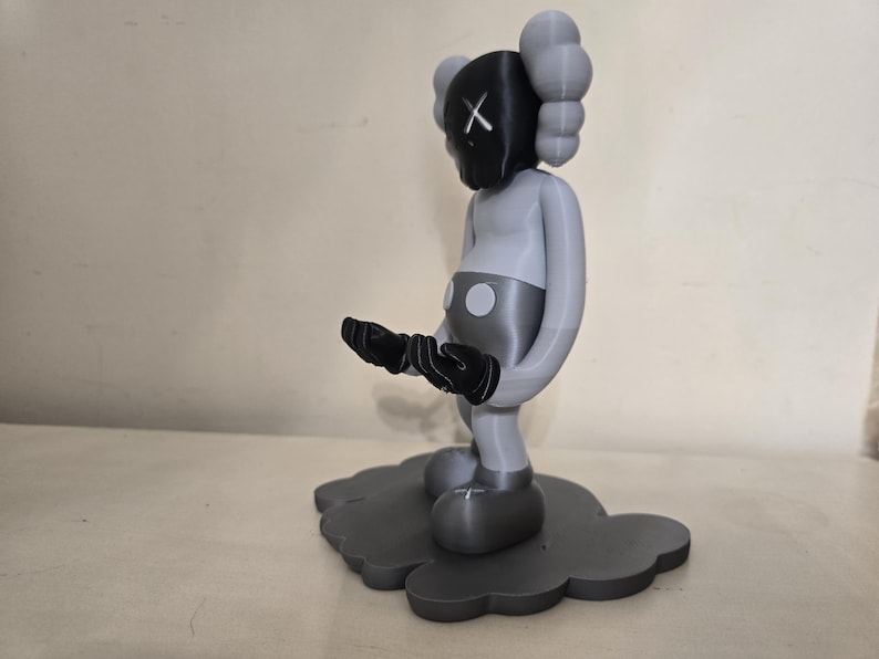 May include: A gray and black figurine of a character with a black X on its face, standing on a gray cloud-shaped base.
