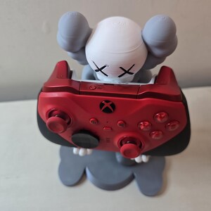 May include: A red Xbox controller is held by a gray and white figurine with an X for eyes. The figurine is standing on a gray base.