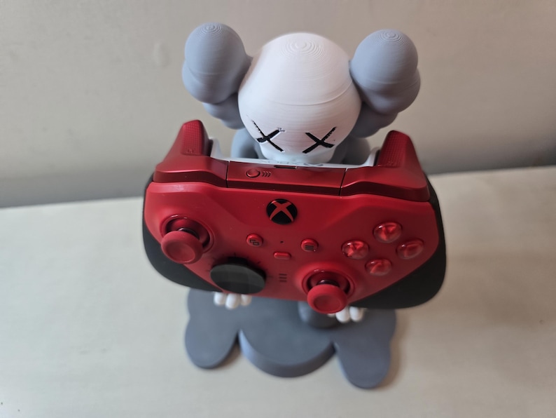 May include: A red Xbox controller is held by a gray and white figurine with an X for eyes. The figurine is standing on a gray base.
