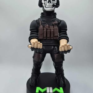 May include: A black figurine of a character with a skull mask and headset, wearing a black tactical outfit. The figurine stands on a black base with the letters "MWII" in green. A skull logo is also on the base.