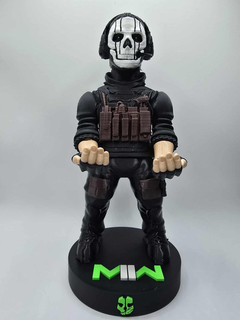 May include: A black figurine of a character with a skull mask and headset, wearing a black tactical outfit. The figurine stands on a black base with the letters "MWII" in green. A skull logo is also on the base.