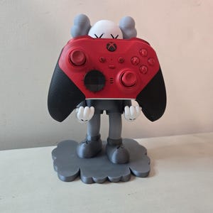 May include: A gray and white figurine holding a red Xbox controller. The figurine is standing on a gray cloud-shaped base.