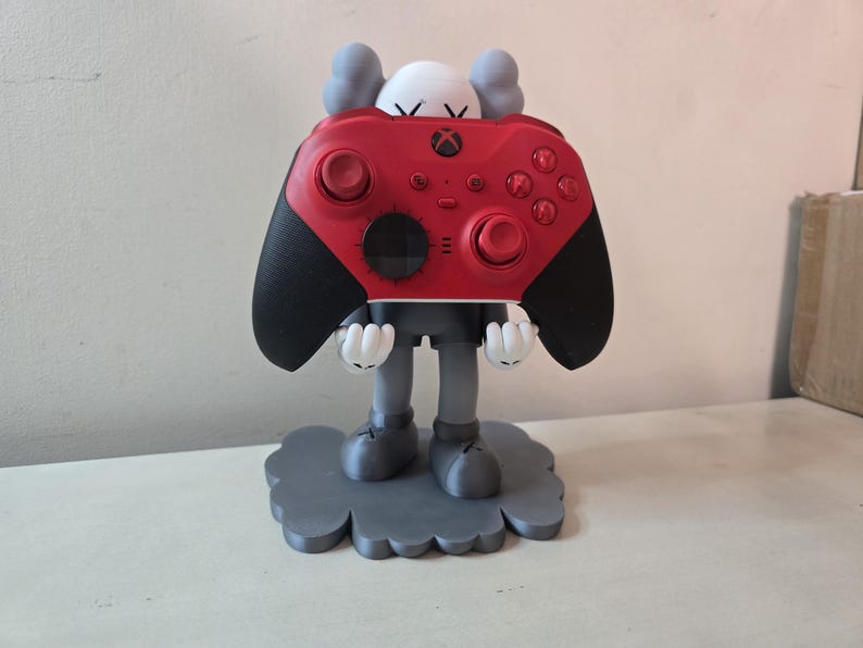 May include: A gray and white figurine holding a red Xbox controller. The figurine is standing on a gray cloud-shaped base.