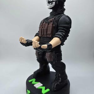 May include: A black figurine of a character in tactical gear, featuring a skull mask and headset. The figure has a brown vest and is standing on a black base with the letters "MW" in green and white.