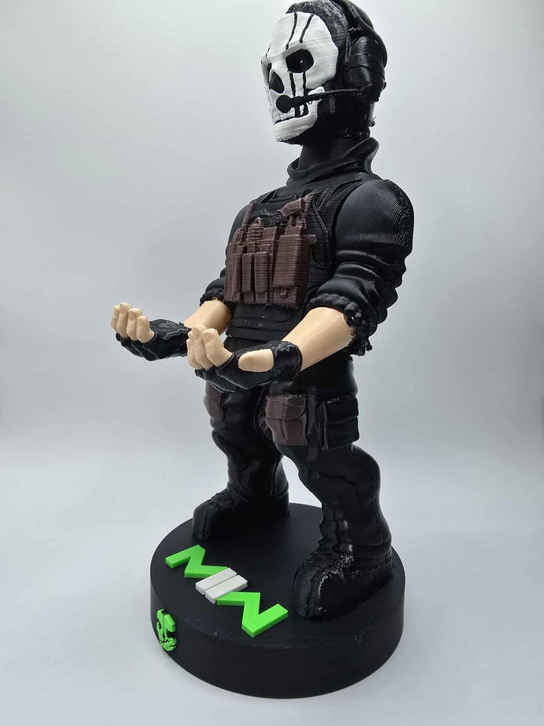 May include: A black figurine of a character in tactical gear, featuring a skull mask and headset. The figure has a brown vest and is standing on a black base with the letters "MW" in green and white.