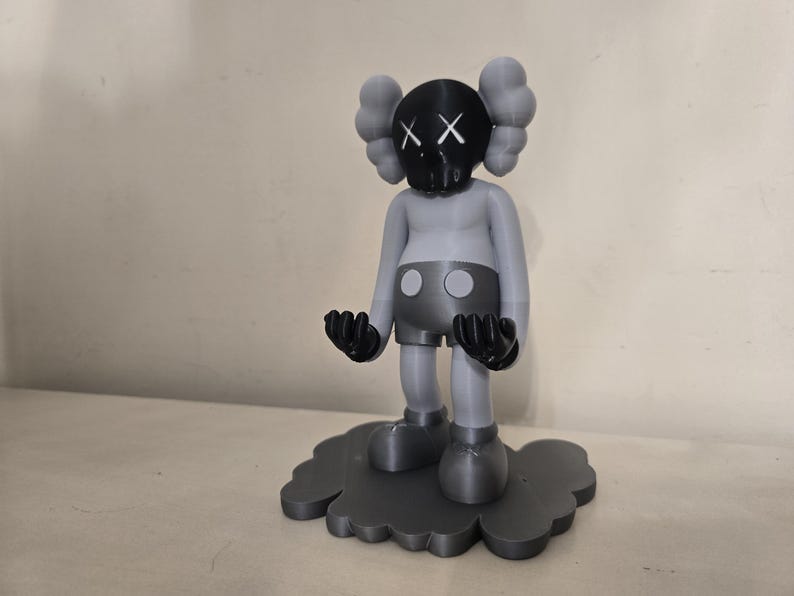 May include: A gray and black figurine of a character with large ears and a black face with an 'X' pattern. The figurine is standing on a gray cloud-shaped base.