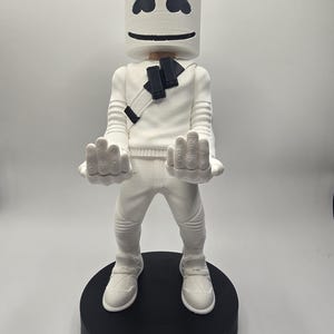 May include: A white figurine of the Marshmello character from Fortnite, with black accents on the eyes, mouth, and chest. The figure is standing on a black base with the word "FORTNITE" in white.