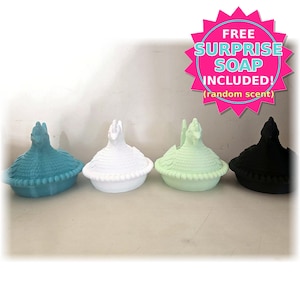 May include: Four chicken-shaped lidded dishes in teal, white, light green, and black. A pink starburst graphic in the upper right corner contains the text: FREE SURPRISE SOAP INCLUDED! (random scent). Each dish features a textured, feathered lid.