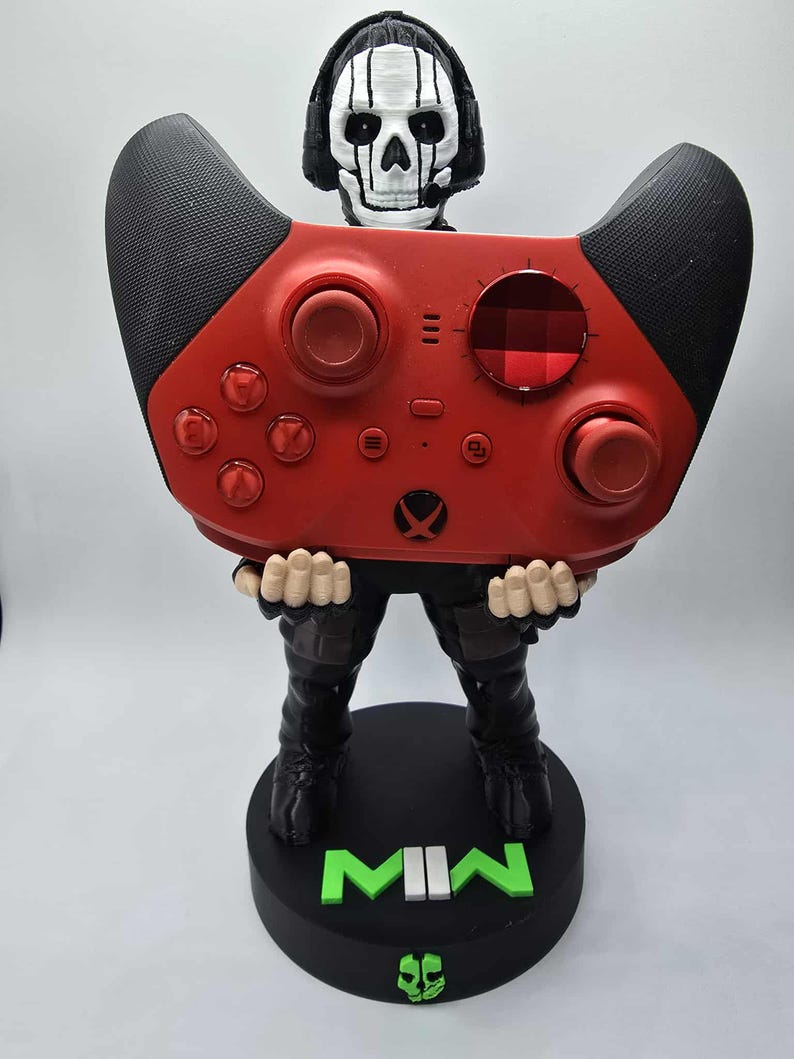May include: A red and black Xbox controller held by a figurine of a skull-faced character wearing a headset. The figurine is standing on a black base with the letters "MWII" in green. The controller has red buttons and a black textured grip.