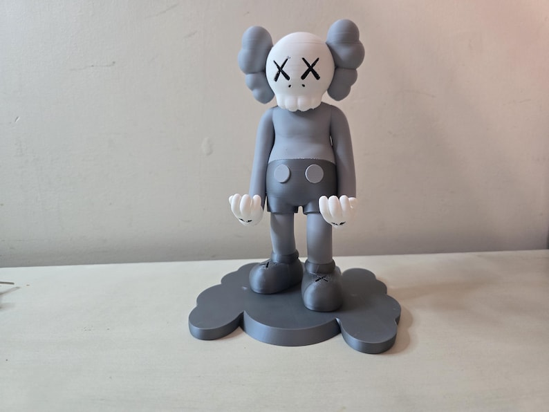 May include: A gray and white figurine of a character with a skull-like head and the letters "XX" for eyes. The figurine is standing on a gray cloud-shaped base.