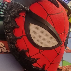 May include: A red and black Spider-Man mask with a black, textured, spiky design around the edges. The mask has a white mesh eye piece.
