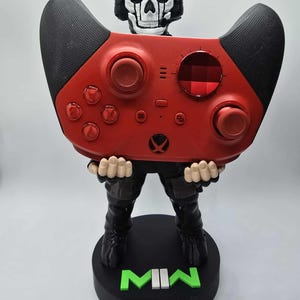 May include: A red and black video game controller held by a figurine of a skull-faced character wearing a headset. The figurine is standing on a black base with the letters "MW" in green.