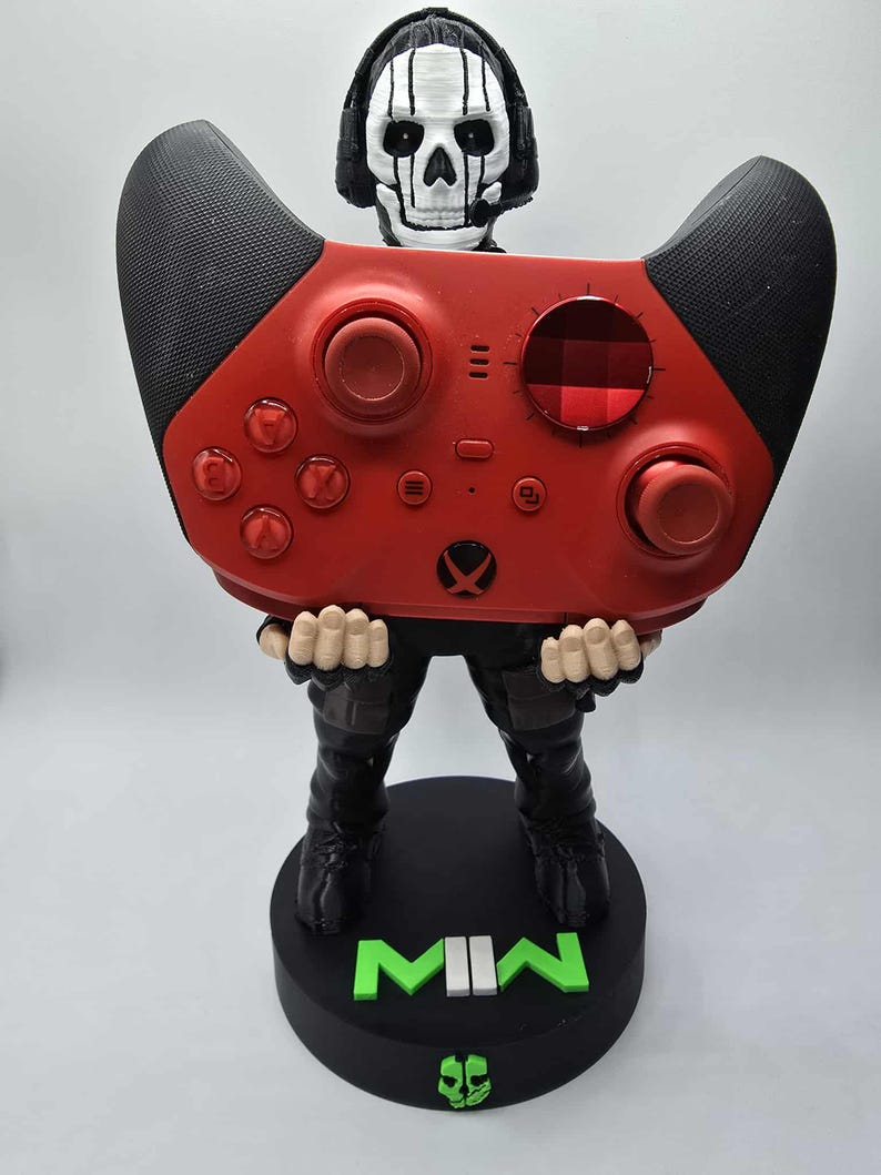 May include: A red and black video game controller held by a figurine of a skull-faced character wearing a headset. The figurine is standing on a black base with the letters "MW" in green.