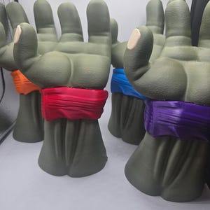 May include: A collection of four green, three-fingered hand sculptures, each featuring a distinct colored wristband in red, orange, blue, and purple. The hands are textured with a matte finish and are positioned upright in a group.