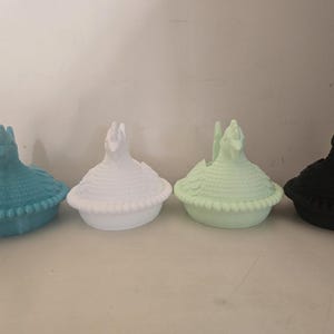 May include: Four decorative lidded containers in a row, each with a bird-like finial. The containers are in shades of teal, white, light green, and black. The lids have a textured, feathered appearance, and the bases have a beaded trim.