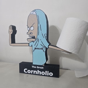 May include: A novelty toilet paper holder featuring a cartoon character in a blue shirt with the word "METALLICA" and the text "The Great Cornholio" in white on a black base. A roll of white toilet paper is attached.