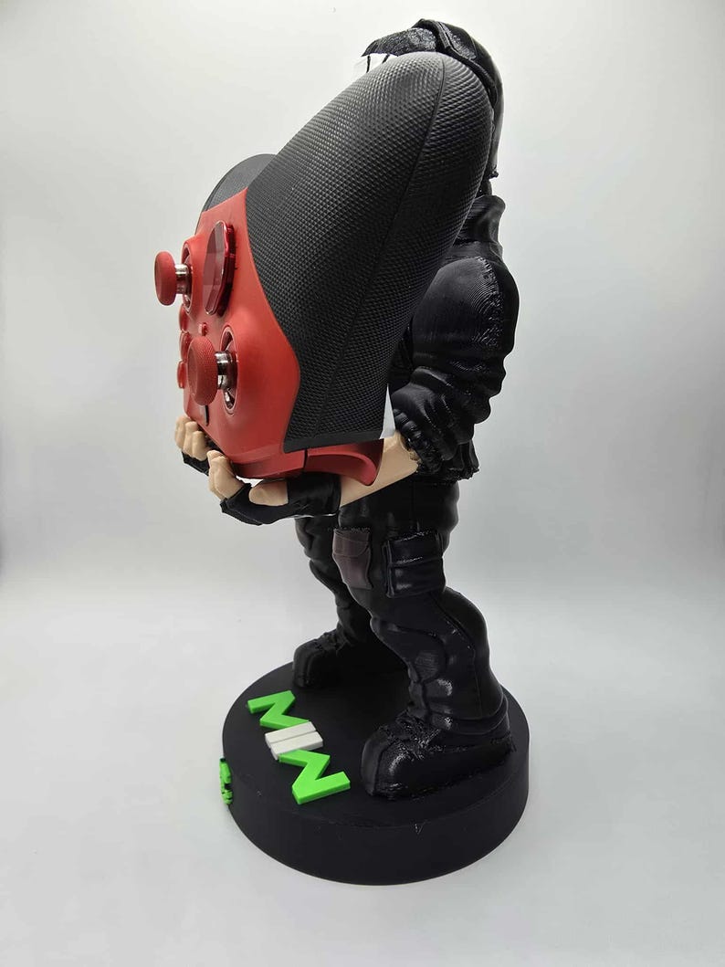 May include: A black and red gaming controller stand featuring a figure in a black outfit. The controller is red with black grips. The base is black with green text that reads "MWN".