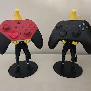 May include: Two 3D printed figures holding video game controllers. The figures are wearing black pants and have yellow hair. One controller is red and the other is black.