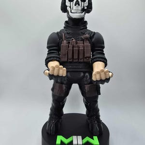 May include: A black figurine of a character with a skull mask and headset, wearing tactical gear. The figure stands on a black base with the letters "MIN" in green and white. A green skull logo is at the base.