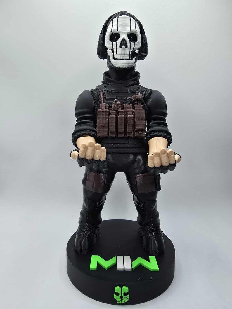 May include: A black figurine of a character with a skull mask and headset, wearing tactical gear. The figure stands on a black base with the letters "MIN" in green and white. A green skull logo is at the base.