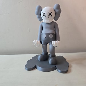 May include: Gray and white figurine of a character with a skull-like head and large, round eyes. The character is wearing a gray shirt and shorts with black buttons. The figurine is standing on a gray cloud-shaped base.